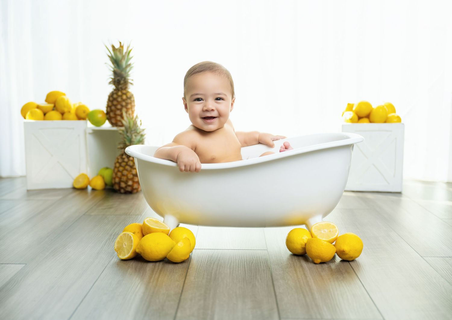 Smiling baby sitting in a white mini bathtub surrounded by lemons and a pineapple on a wooden floor.