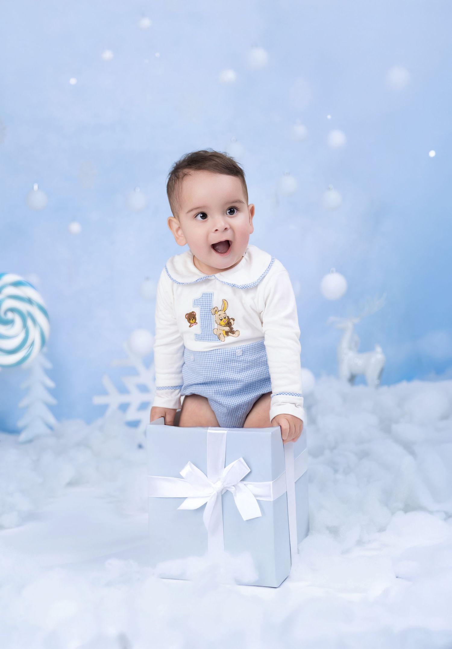 Cheerful baby in white top and blue shorts sits in a gift box amid a winter wonderland holiday backdrop.
