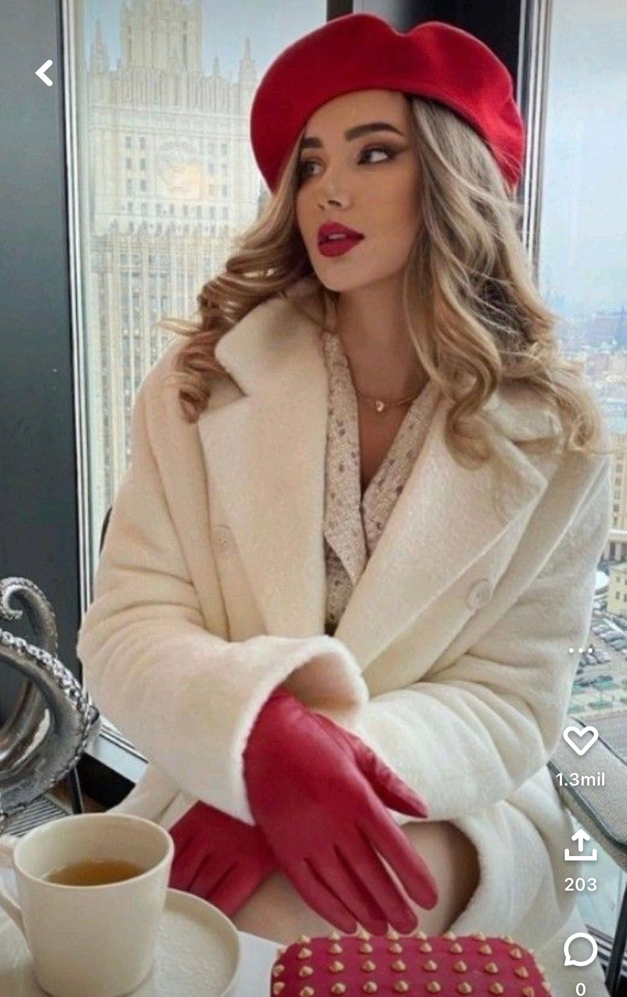 Elegant blonde woman in red beret and gloves holding tea cup, wearing cream coat, seated by city window.
