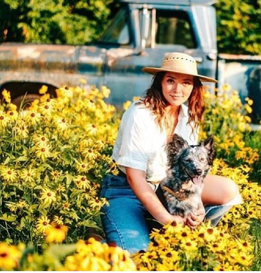 Woman in straw hat sitting among yellow wildflowers holding a dog, with a vintage blue truck in the background.