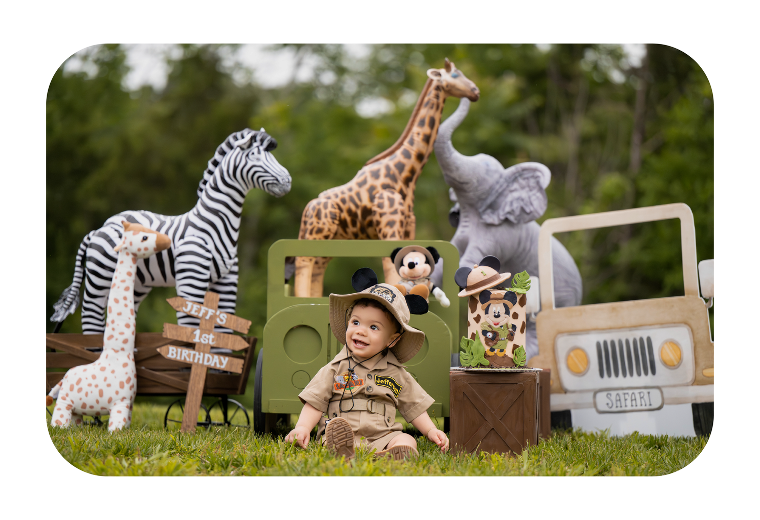 Baby in safari outfit surrounded by stuffed animal toys including giraffe, zebra, and elephant on green grass.
