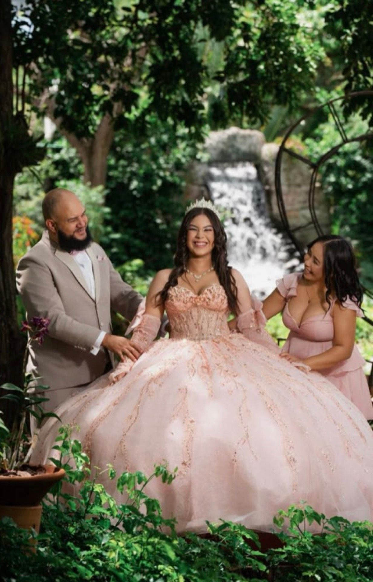 A young woman in a pink quinceañera ball gown with tiara poses with two guests near a waterfall garden backdrop.