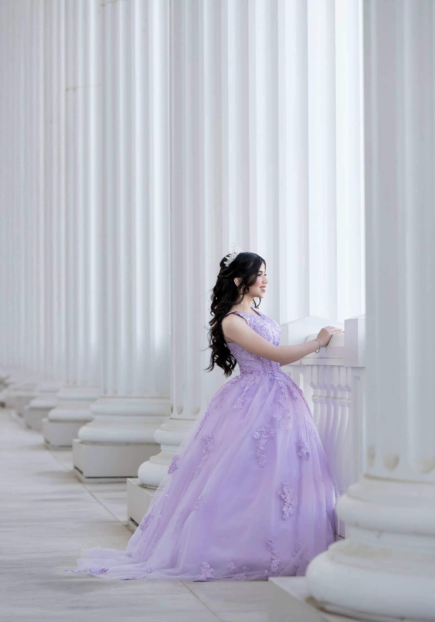 Young woman in lavender ball gown with floral details poses gracefully among white classical columns.