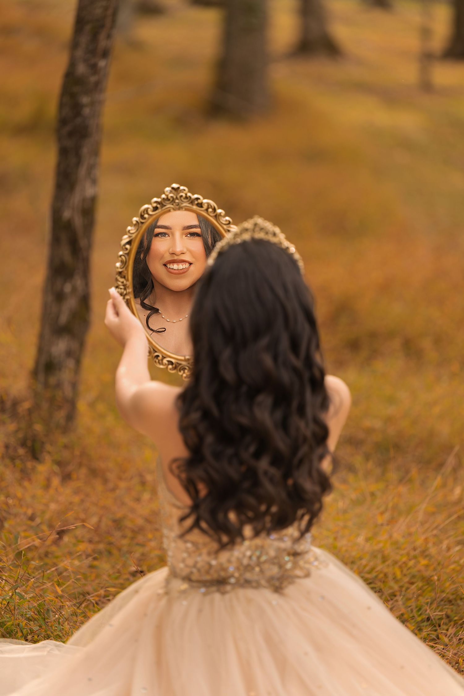 A smiling bride in a champagne gown holds a gold ornate mirror outdoors in an autumn forest setting.