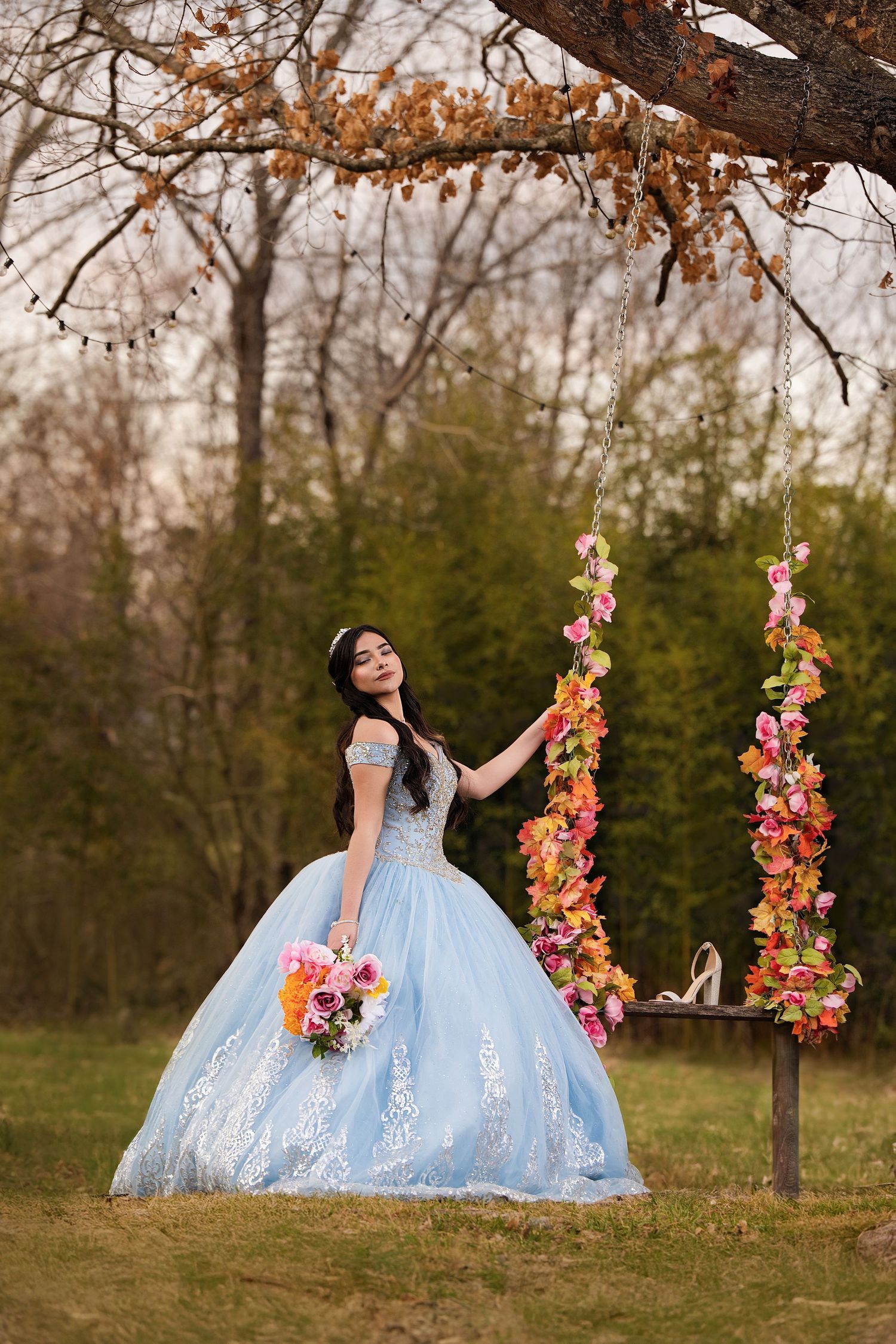 Young woman in blue ball gown holding flowers by a floral swing in an autumn garden setting.