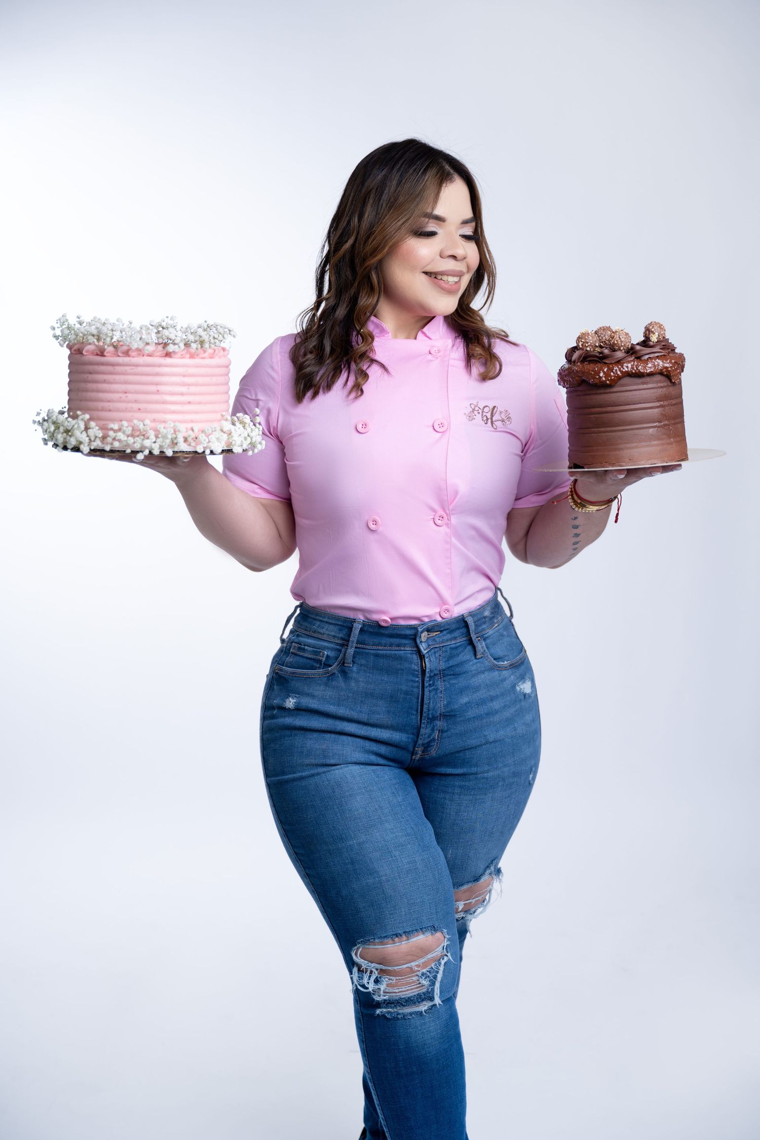 Baker holding pink and chocolate layer cakes while wearing pink chef coat and distressed jeans.