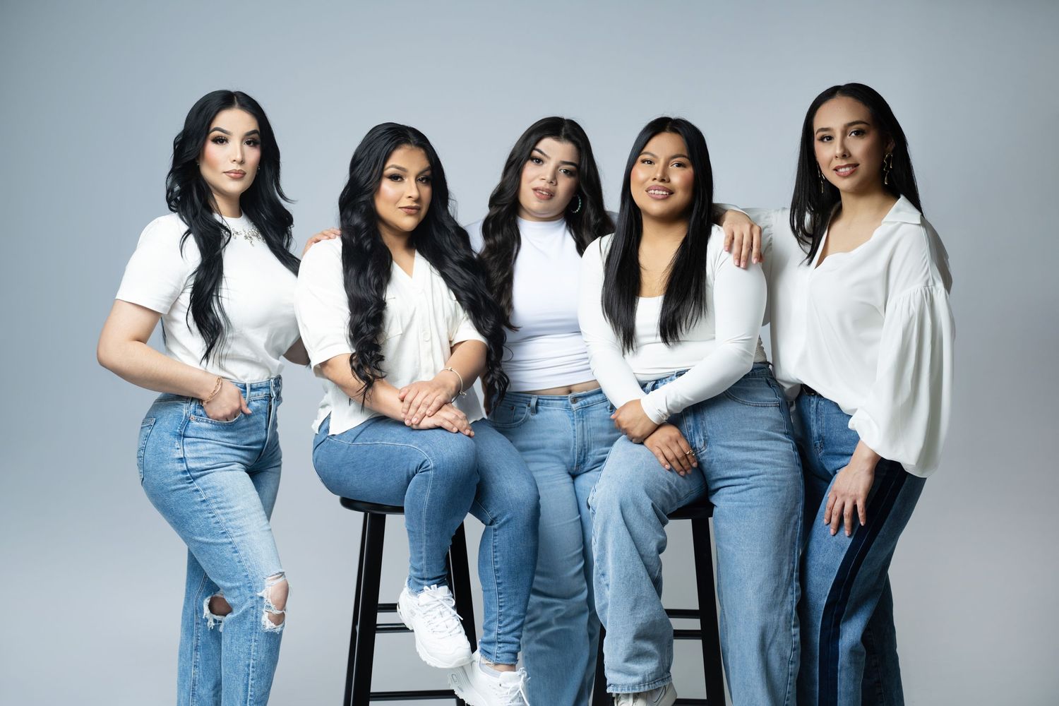 Group of five people in white tops and jeans sitting together on stools against a gray background.