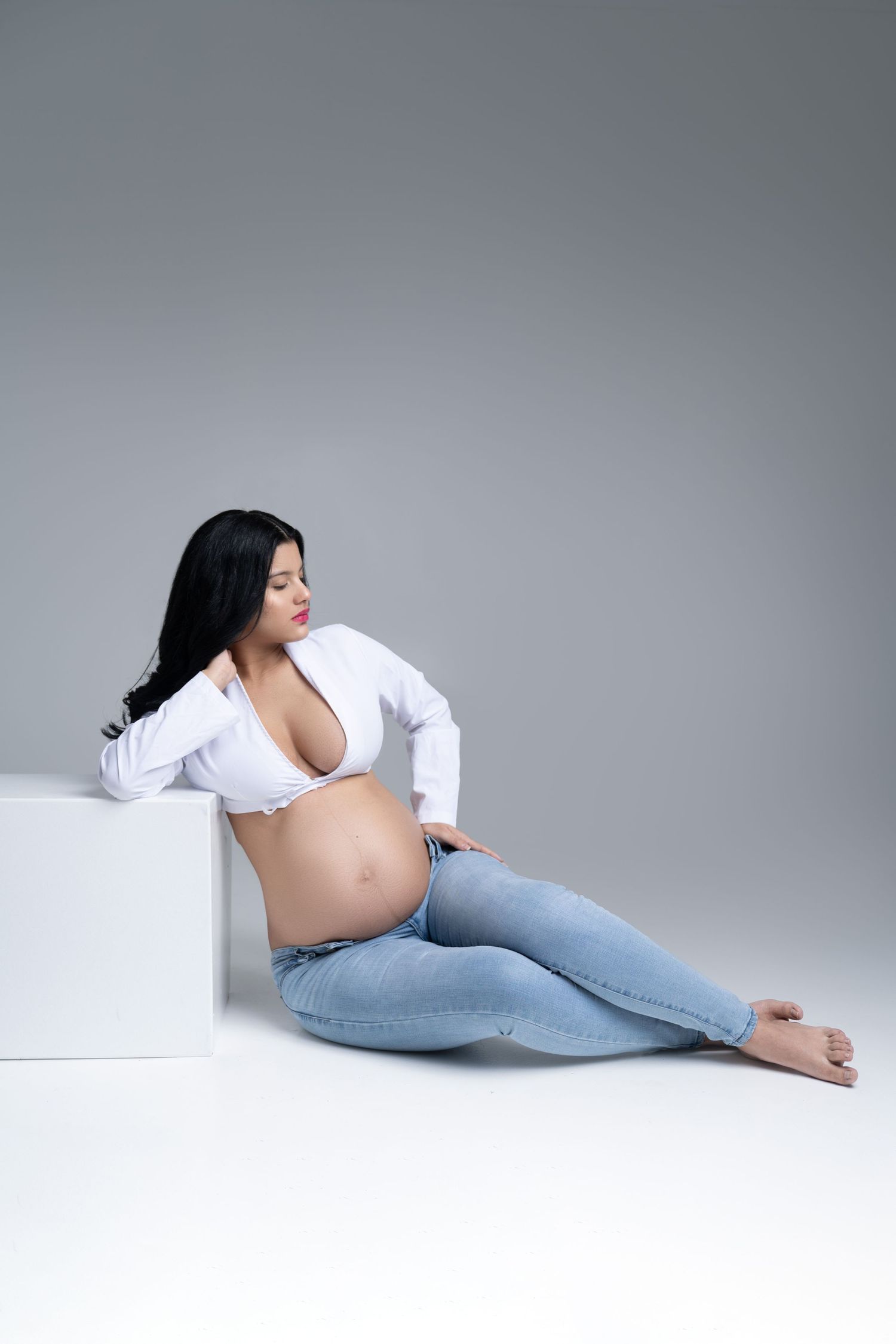 Casual maternity photo with subject in white button-down shirt and jeans lounging against white cube.