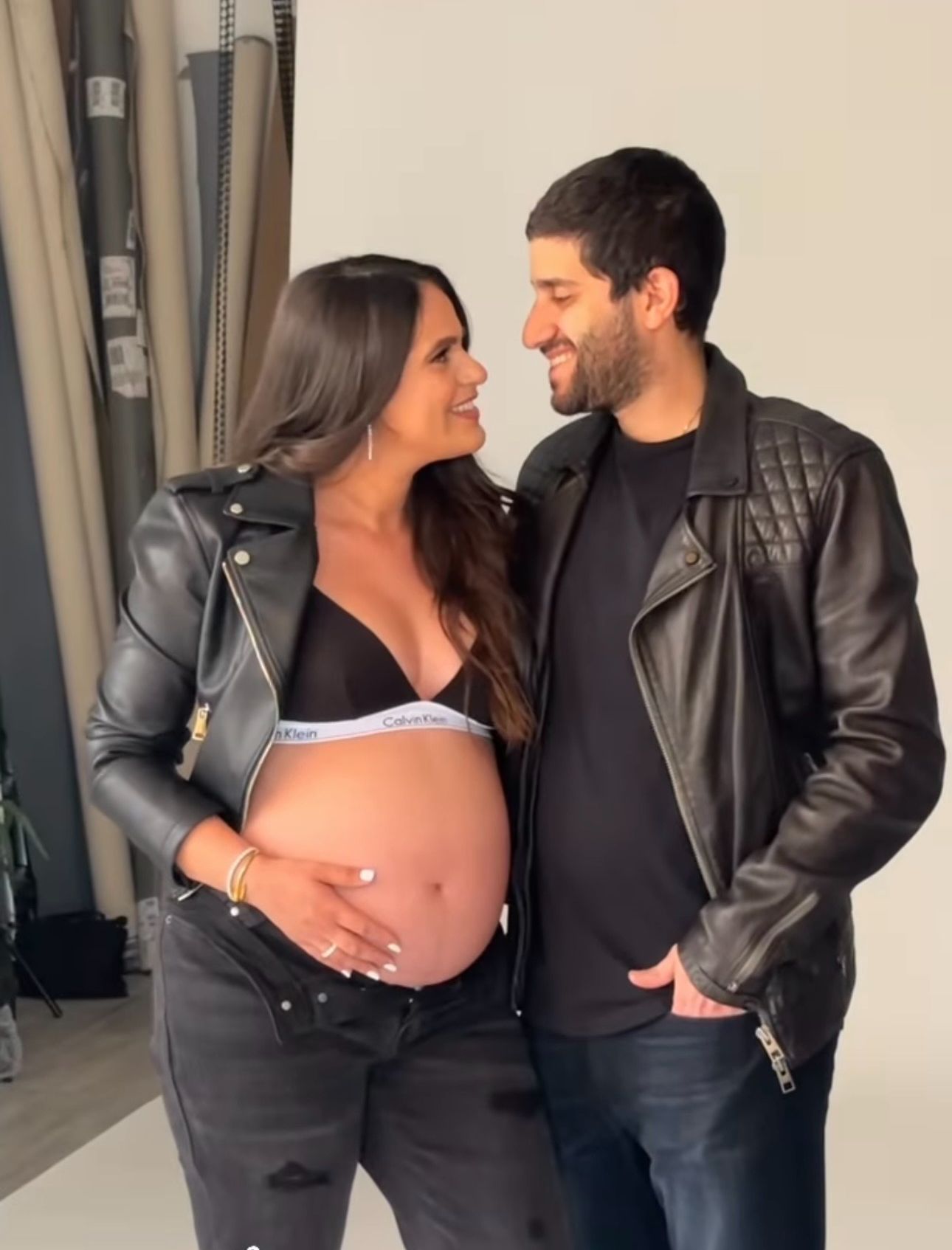 Pregnant woman in Calvin Klein bra and leather jacket poses with smiling man in black leather jacket during maternity photoshoot.