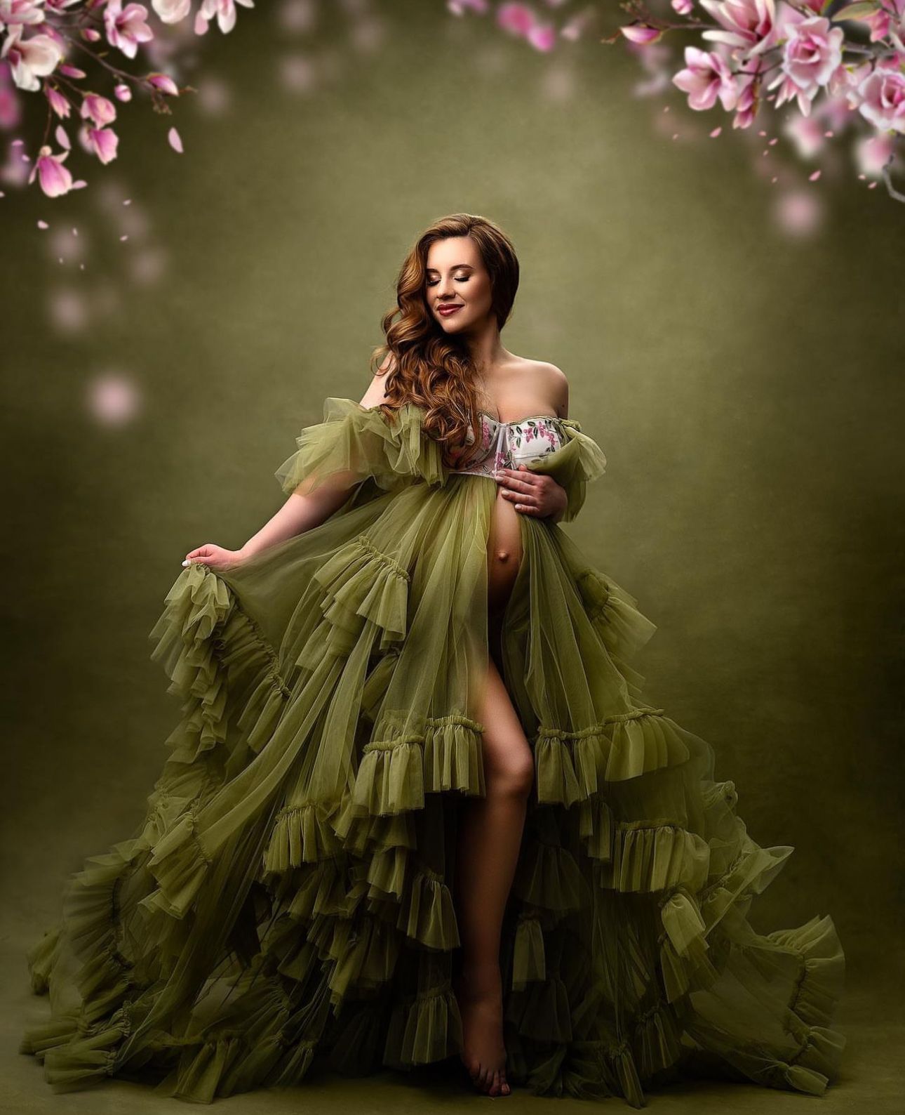 A pregnant woman in an olive green ruffled tulle gown poses gracefully under pink floral branches on a green backdrop.