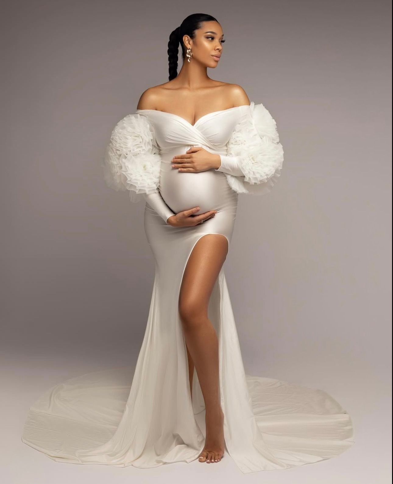Pregnant woman in elegant white off-shoulder gown with ruffle sleeves and thigh-high slit for maternity photoshoot.
