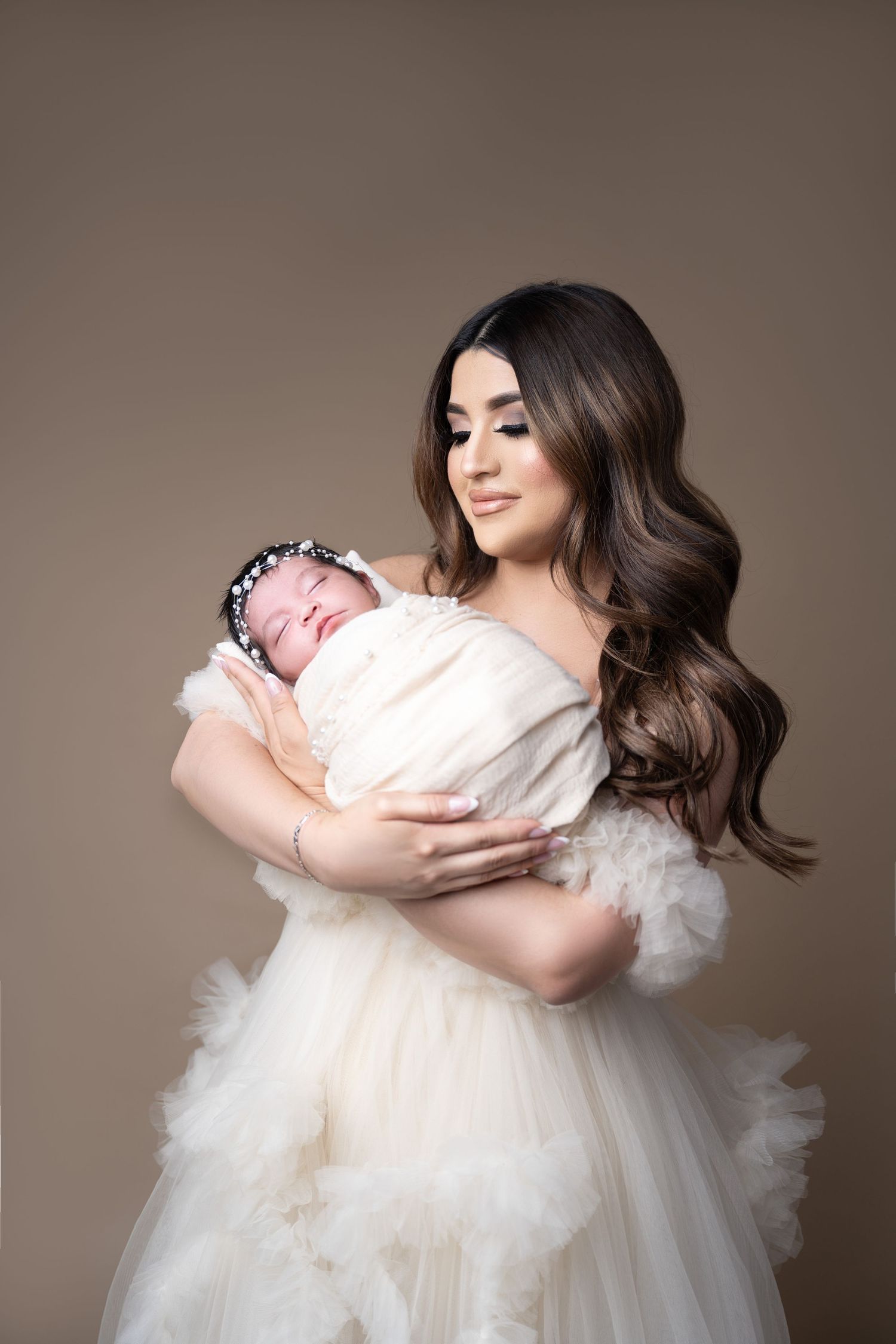 A mother in a white tulle gown tenderly holds her sleeping newborn wrapped in white on a neutral background.