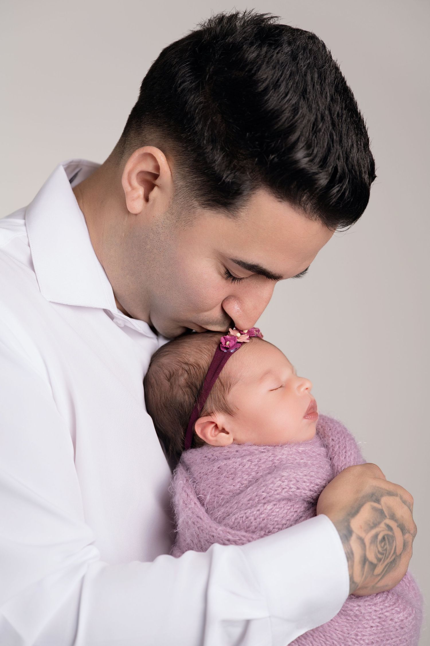 A father in a white shirt gently kisses his sleeping newborn baby girl wrapped in a pink blanket with a floral headband.