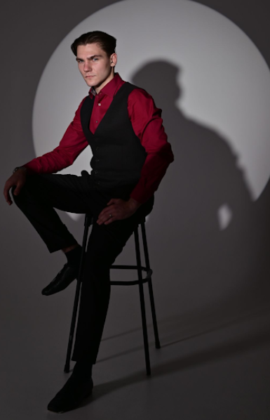 Dramatic studio portraits with person in red shirt and black vest seated on chair against dark background.