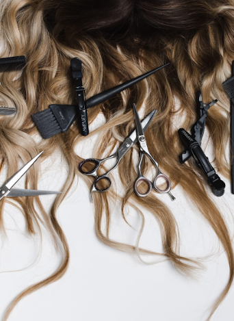 Professional hairstyling tools including scissors, clips and brushes arranged on blonde wavy hair extensions against white background.