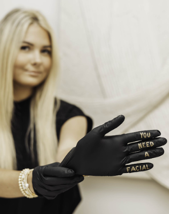 Black gloved hands display gold lettering spelling out 'YOU NEED FACIAL' against a white backdrop.