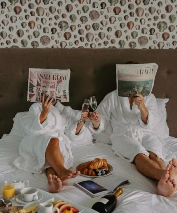 Two people in bathrobes reading magazines in bed with breakfast, orange juice and pastries laid out between them.