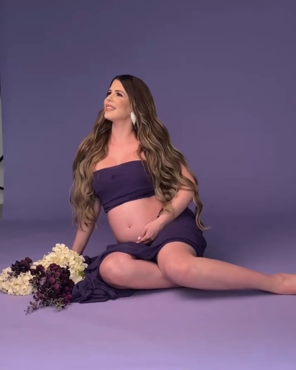 A pregnant woman in purple attire poses gracefully on a purple backdrop with floral arrangements beside her.