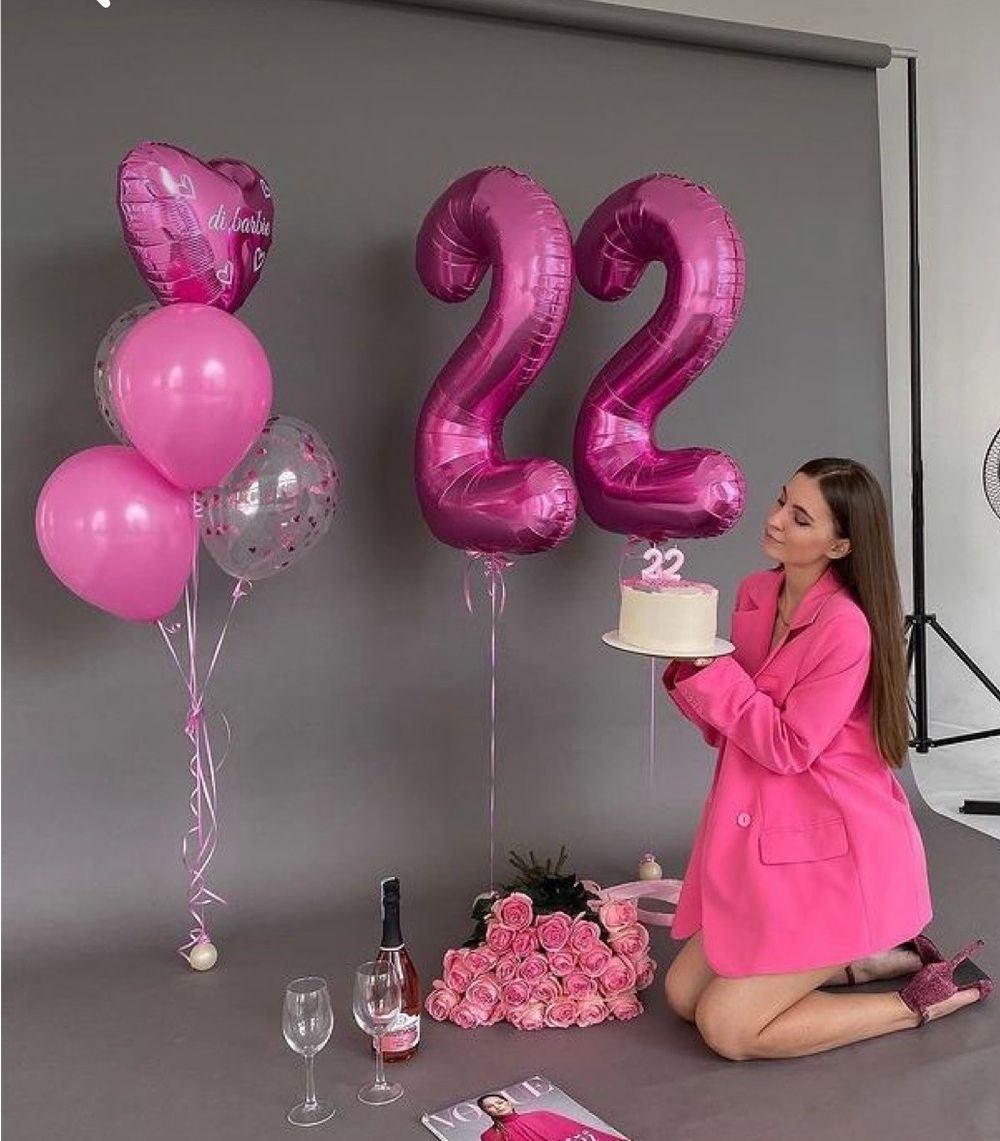 Woman in pink blazer kneeling beside large pink number 22 balloons, holding birthday cake at a pink-themed party.