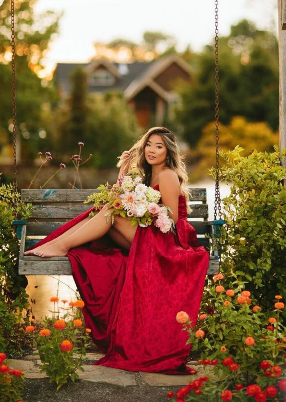 Woman in red gown holding flowers on garden swing surrounded by blooming roses at golden hour.