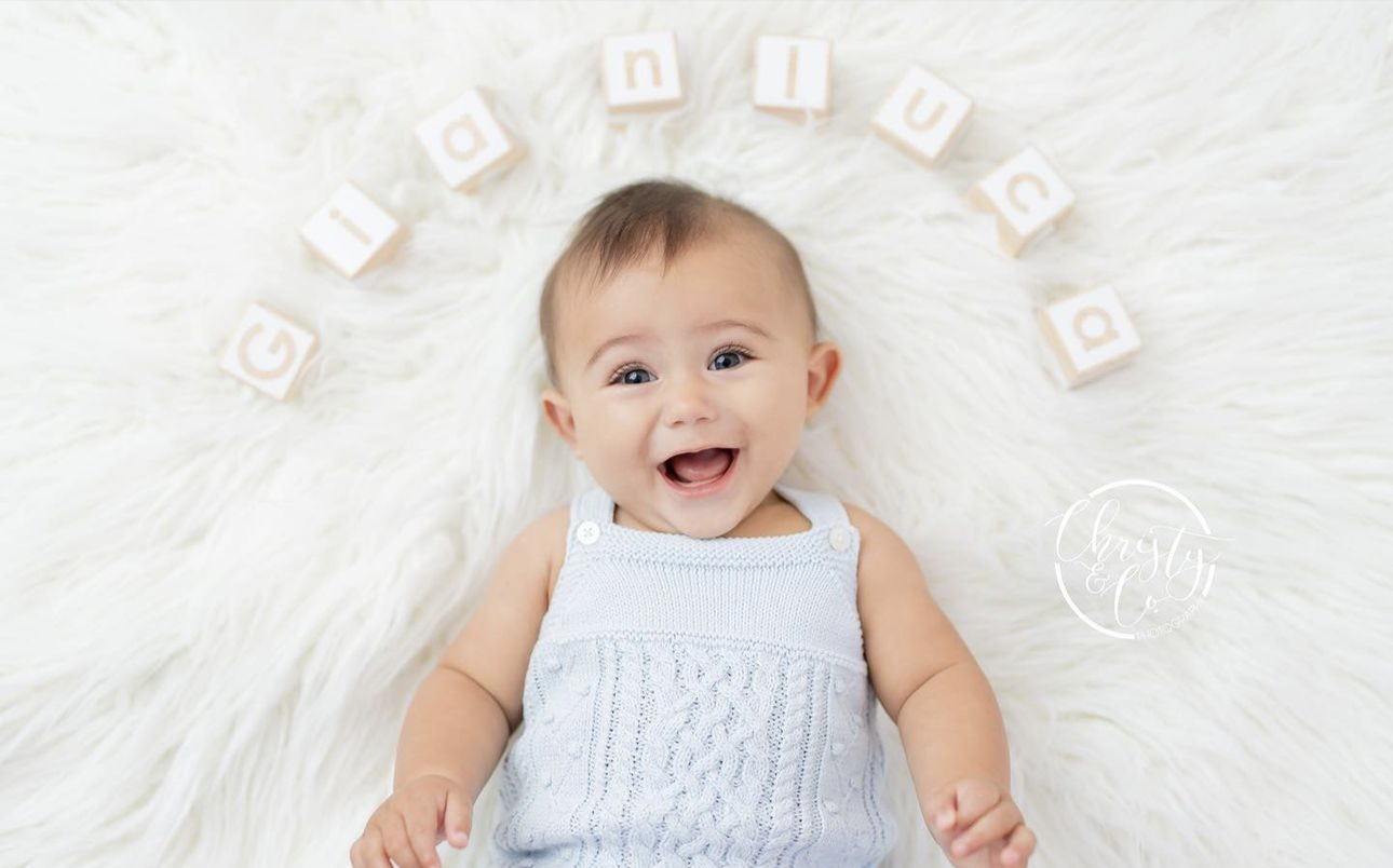 Smiling baby in blue romper lying on white fur rug surrounded by wooden alphabet blocks spelling a name.