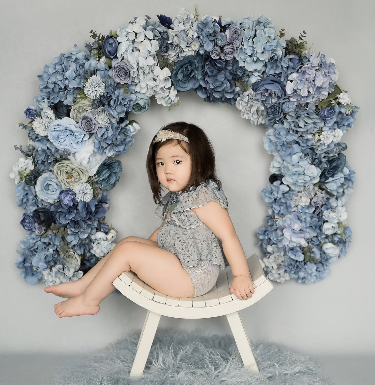 Adorable toddler girl sitting on white chair inside blue hydrangea floral arch in professional studio portrait.