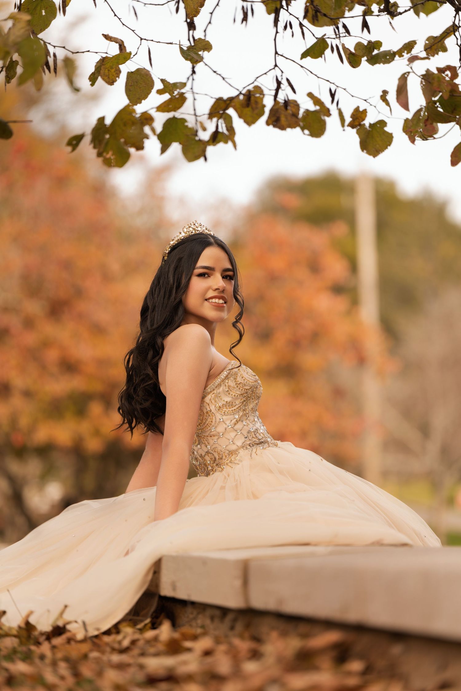 Young woman in gold quinceañera gown and tiara sits outdoors among autumn foliage, smiling gracefully.