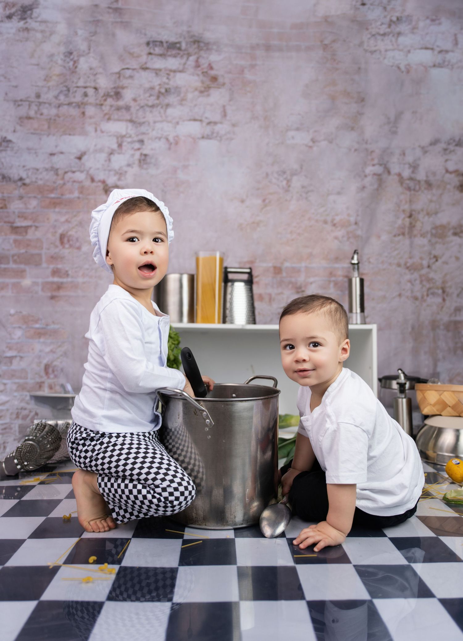 Two toddlers dressed as chefs play with a large pot and spoon on a checkered kitchen floor.