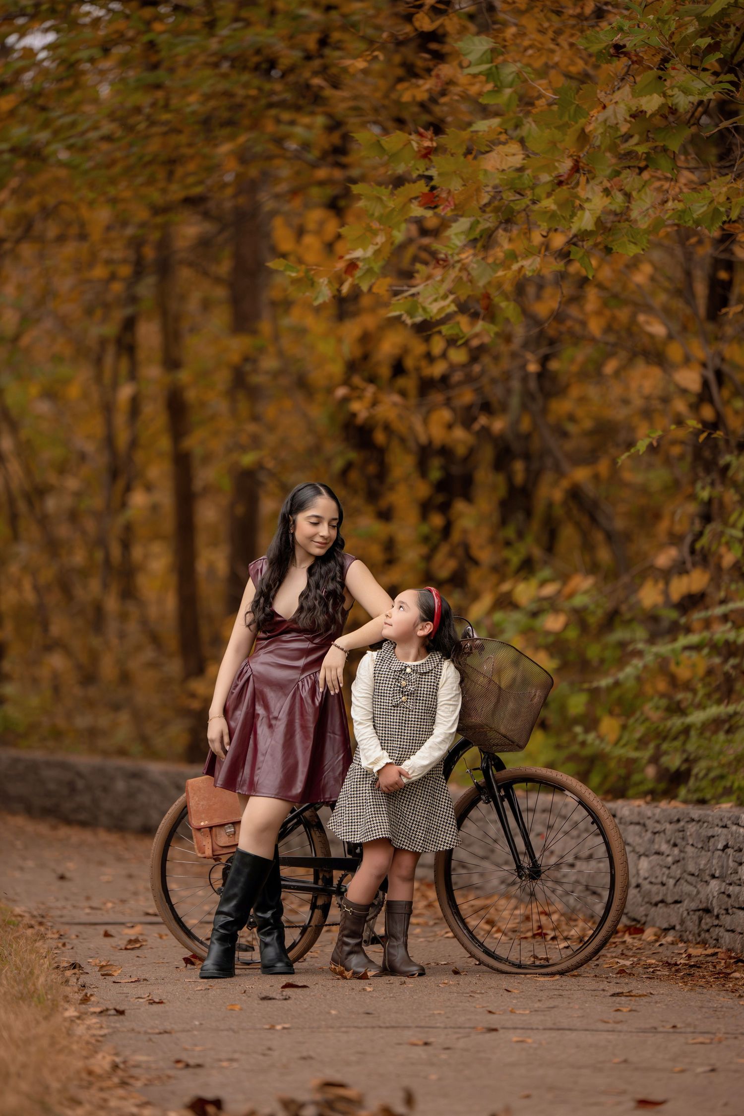 A woman in a burgundy leather dress and black boots poses with a young girl beside a vintage bicycle in an autumn forest.