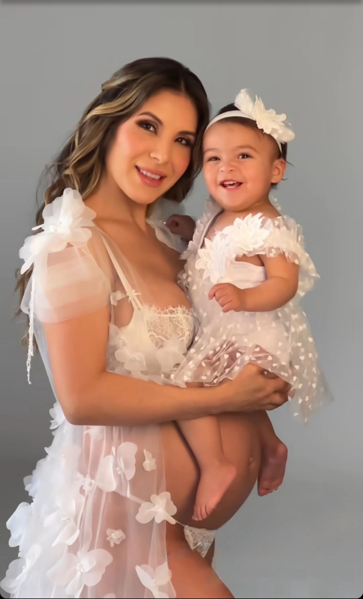A smiling woman in a white floral tulle gown holds a happy baby girl in a matching white dress and floral headband.