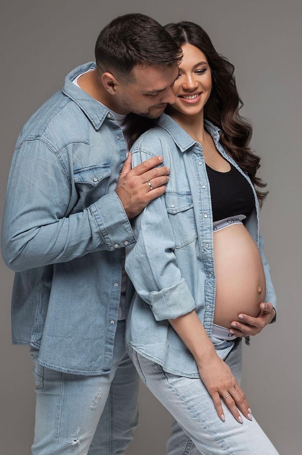 Happy pregnant woman in denim jacket with partner embracing her baby bump in a studio photo shoot.