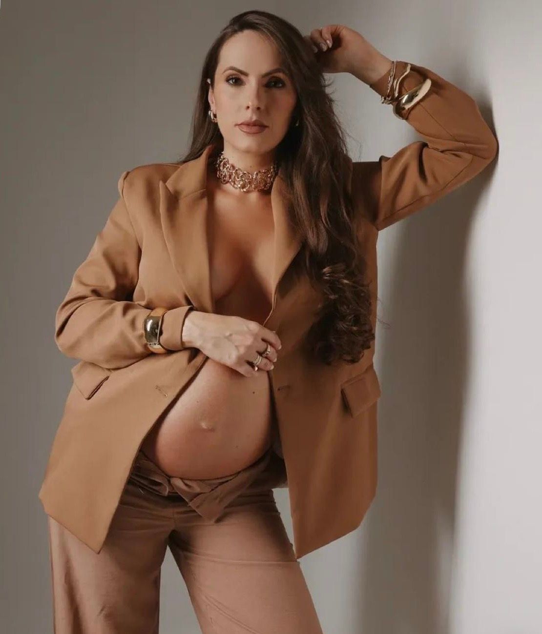 Pregnant woman in tan blazer and pants with gold jewelry posing confidently, showcasing baby bump in stylish maternity look.