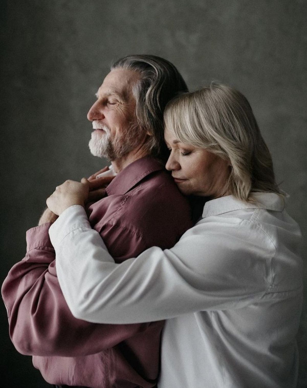 Elderly couple embracing tenderly, woman in white shirt hugging man from behind against gray studio background.