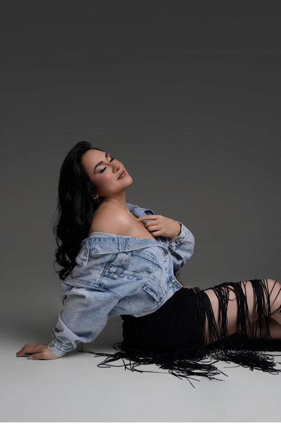 Woman with long dark wavy hair sits on a studio floor, head tilted back, wearing an off-shoulder denim jacket and a black fringe skirt.