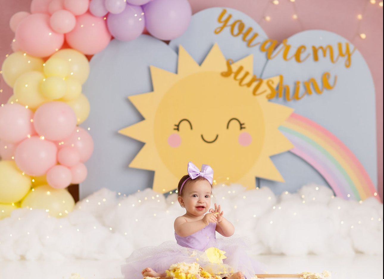 A whimsical cake smash photo setup with pastel balloons, clouds, rainbows and yellow sun decorations against a blue backdrop.