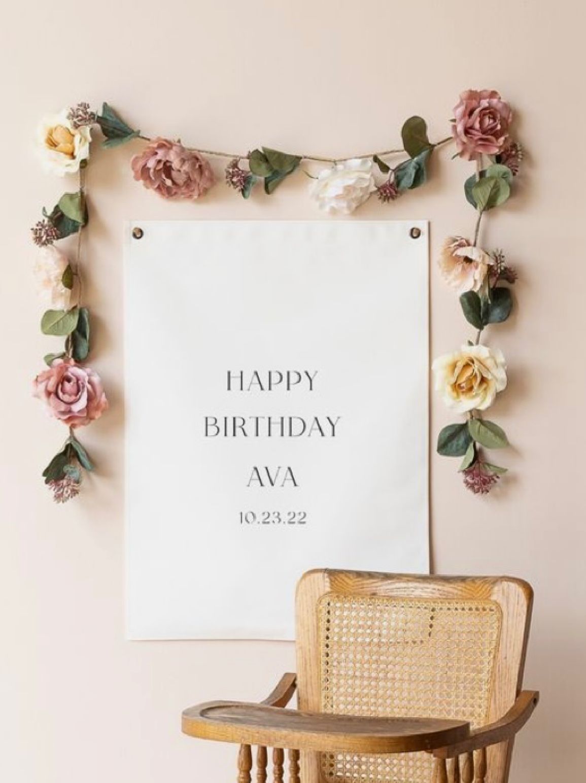 Birthday sign decorated with pink and yellow roses and eucalyptus leaves above a rattan chair.