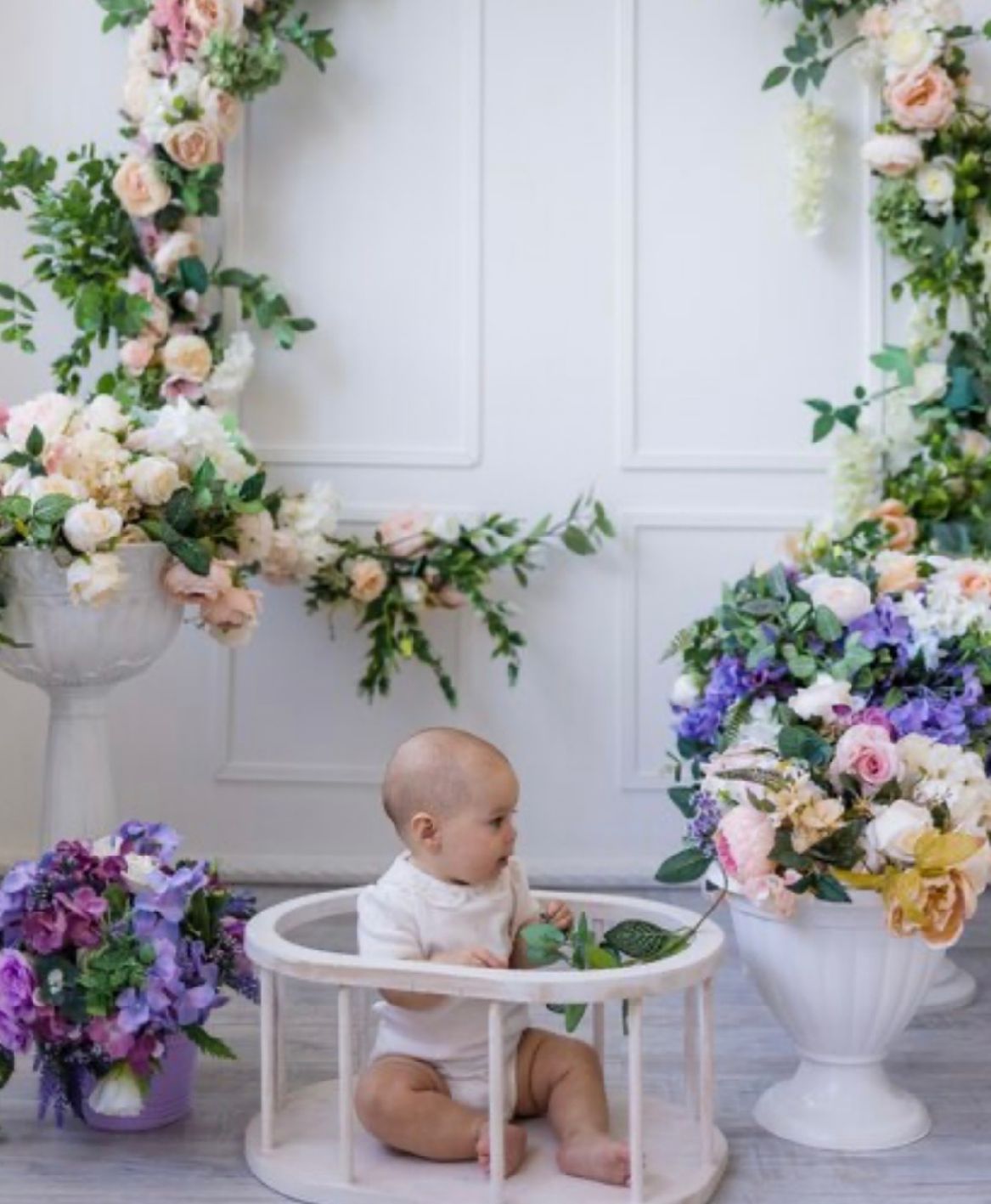 Baby sits in a white circular chair surrounded by floral arrangements and decorative elements for a photo session.