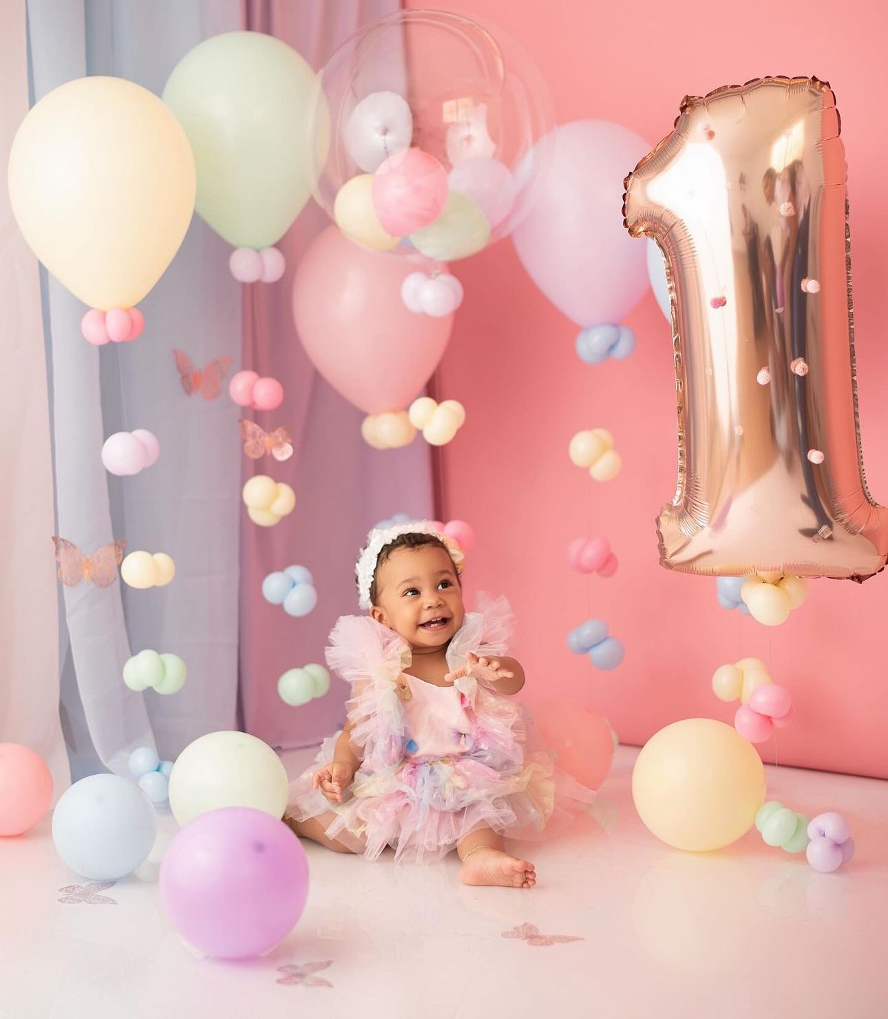 Baby in pink tutu dress sits among pastel balloons and decorations for first birthday celebration.