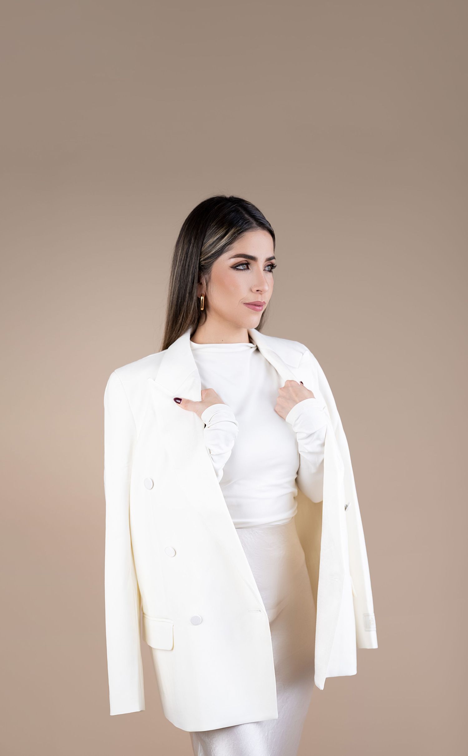 Woman in white blazer and outfit poses confidently against a beige background, showcasing elegant fashion style.