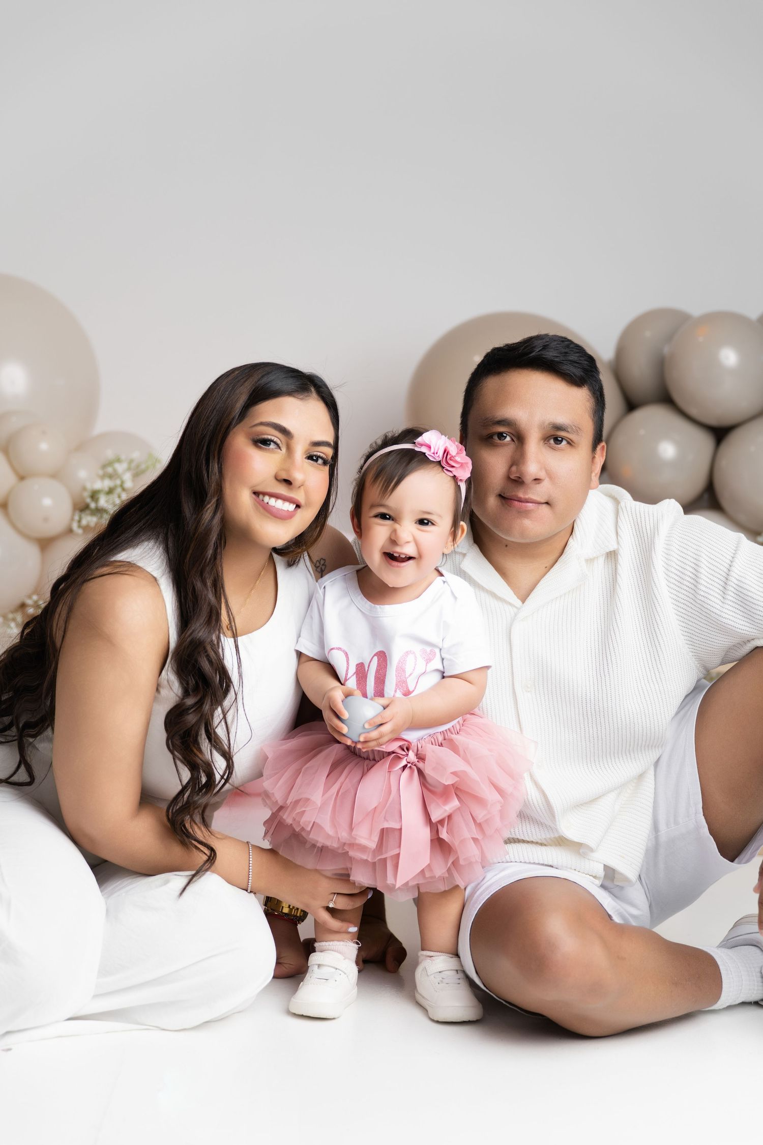 Family of three posing for a first birthday photo with pink tutu baby, balloon backdrop, and parents in white outfits.