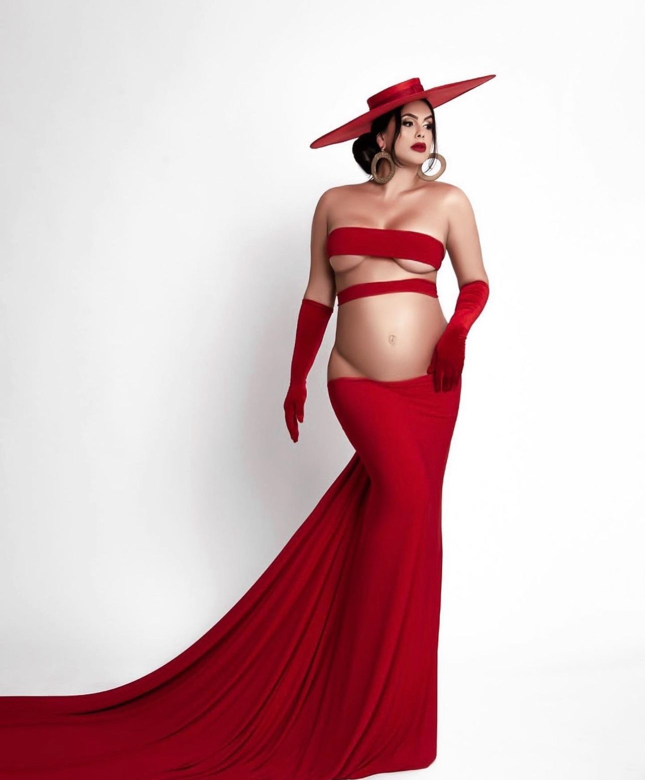 Pregnant woman in red strapless crop top, long gloves, wide-brim hat, and flowing train skirt for a maternity photoshoot.