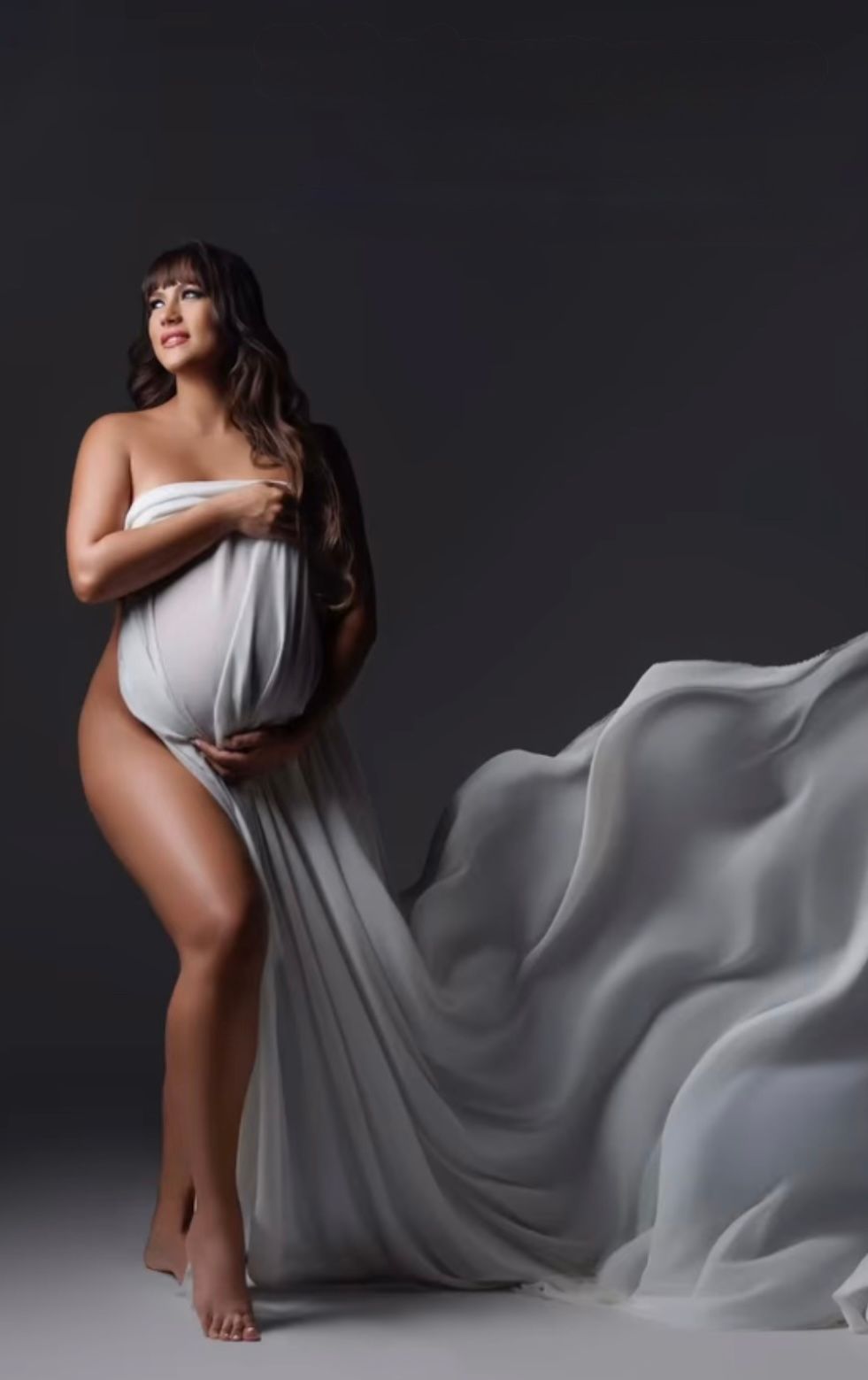 Pregnant woman draped in flowing white fabric poses gracefully in a studio maternity photography session.