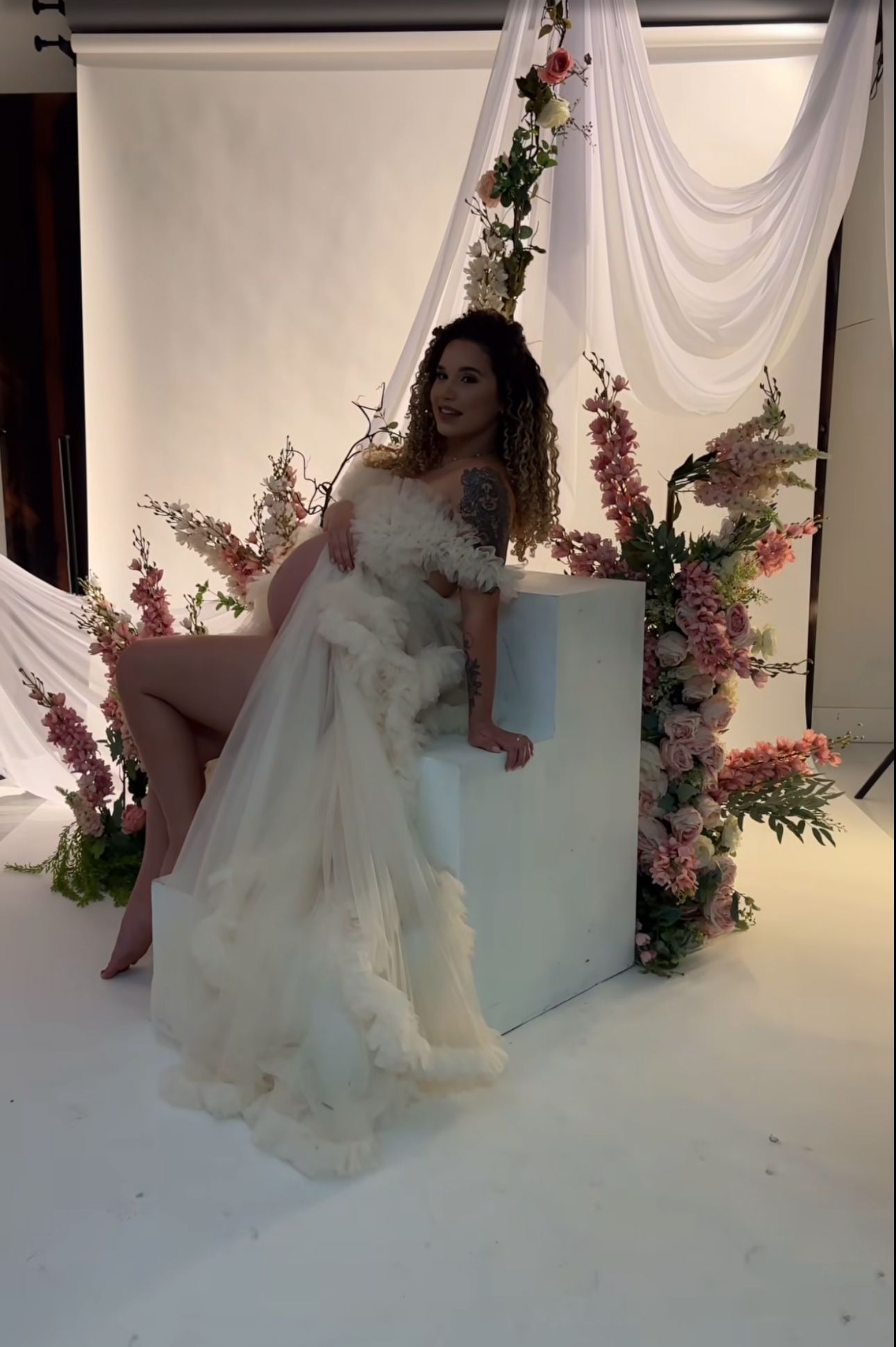 Woman in white ruffled tulle gown sits beside floral arrangement on white box in studio setting.