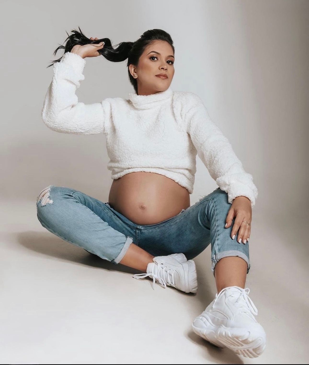 Pregnant woman in white crop sweater and ripped jeans sits on floor holding her hair up, showing baby bump.
