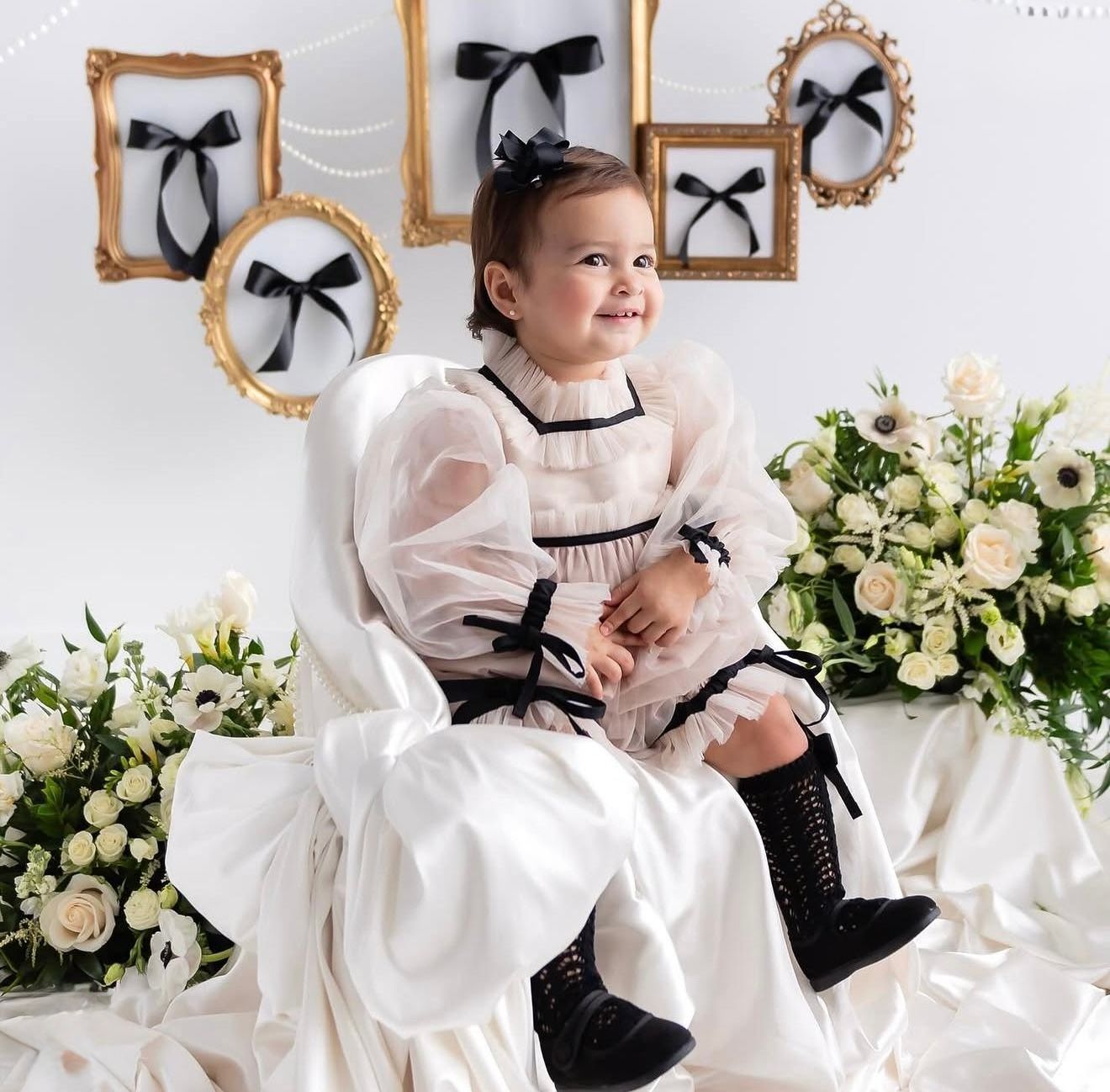 Young girl in white tulle dress with black bows sits on white chair surrounded by white flowers and gold frames.