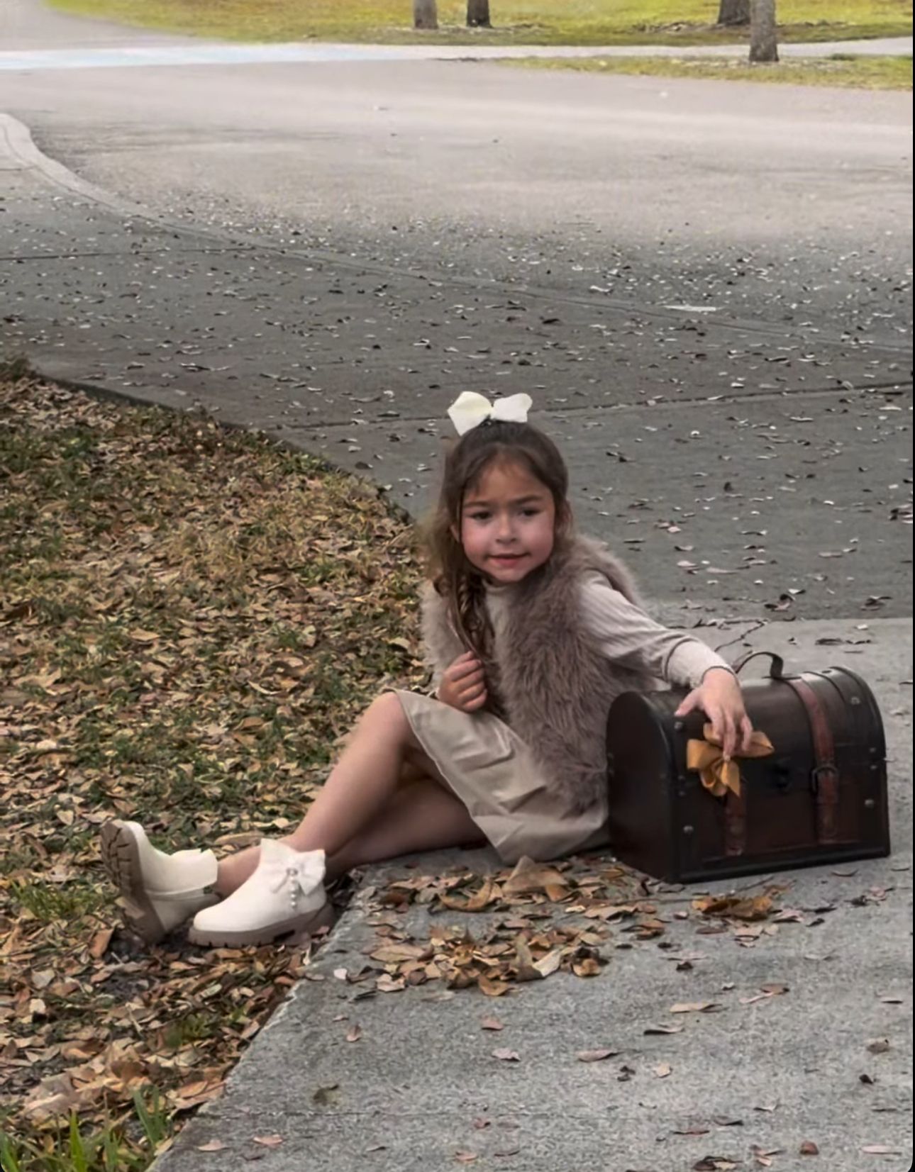 Young girl in fur vest and white boots sits on curb with vintage treasure chest amid autumn leaves.