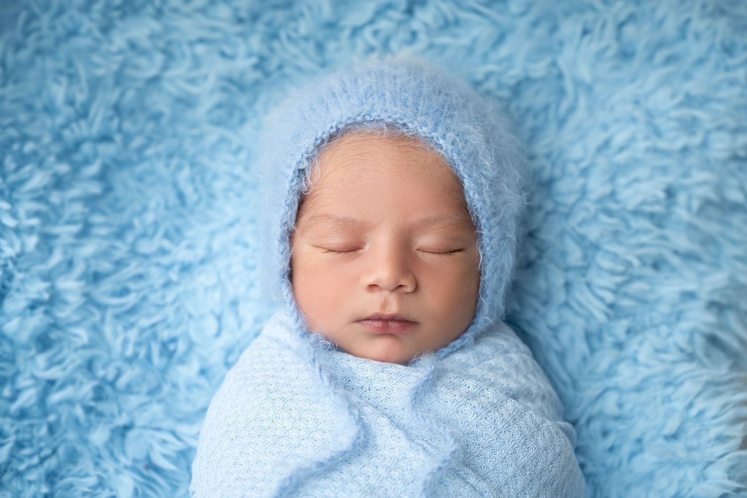 Peaceful sleeping newborn in soft blue knit bonnet and wrap on textured blue background.