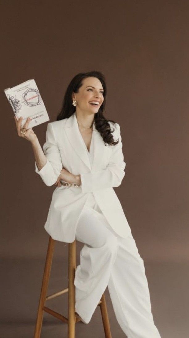 A smiling woman in a white suit sits on a stool holding a book against a brown background.
