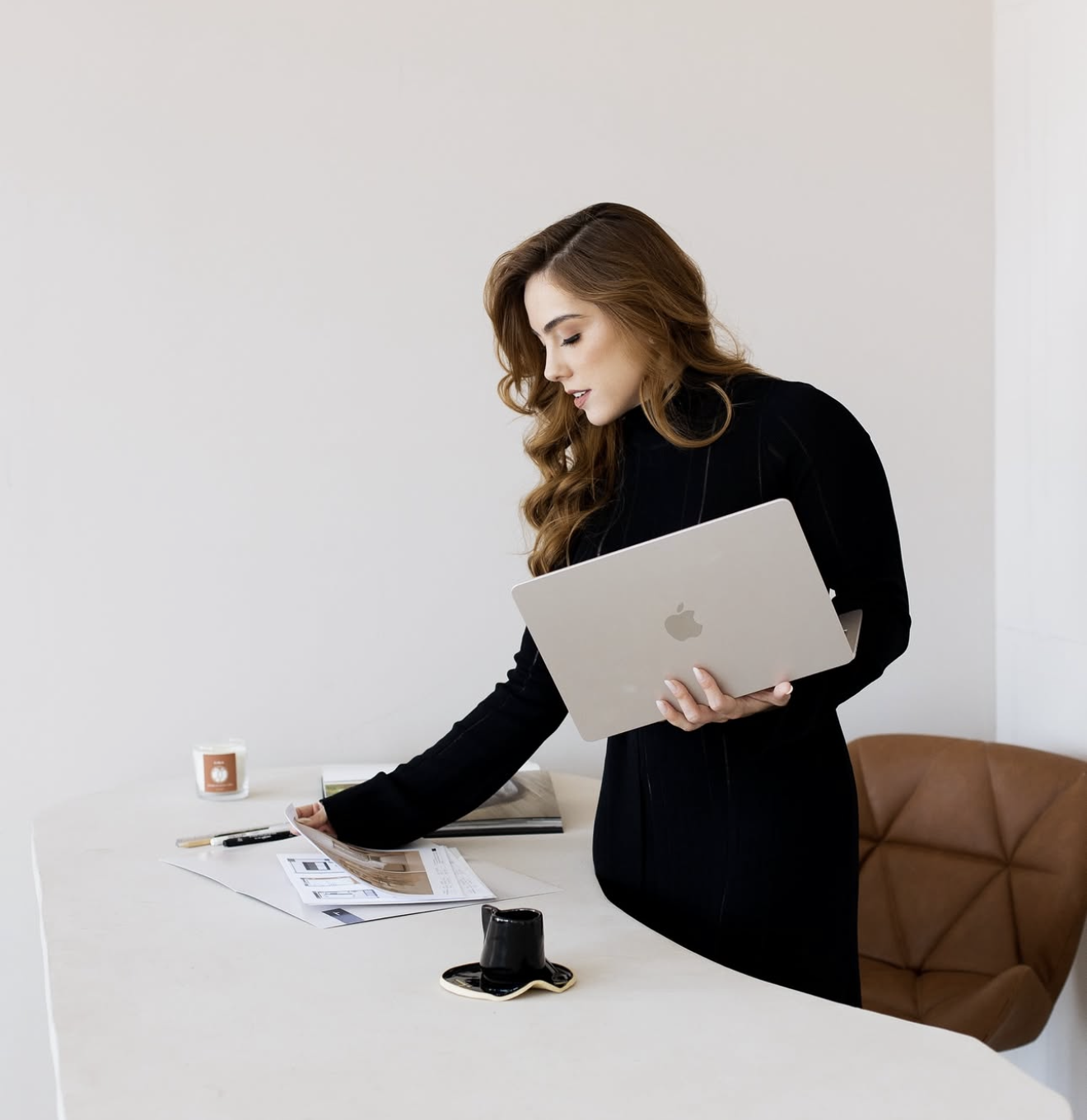 Woman in black dress holding MacBook laptop while standing at a modern white desk in a minimalist office.