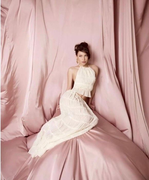 Woman in white halter crop top and skirt posed against flowing pink satin fabric backdrop.